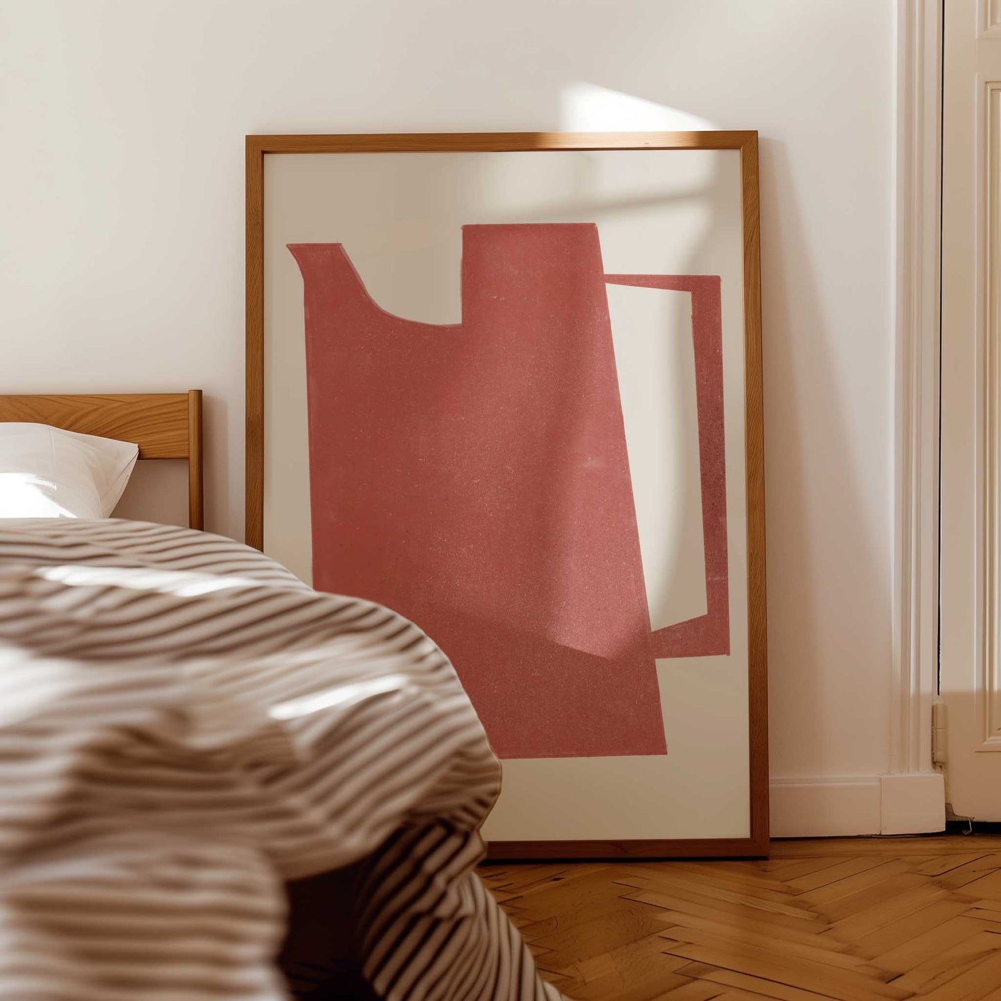 A large framed abstract art print with a terracotta red and white geometric design, leaning against a white wall in a bedroom next to a bed with striped bedding.
