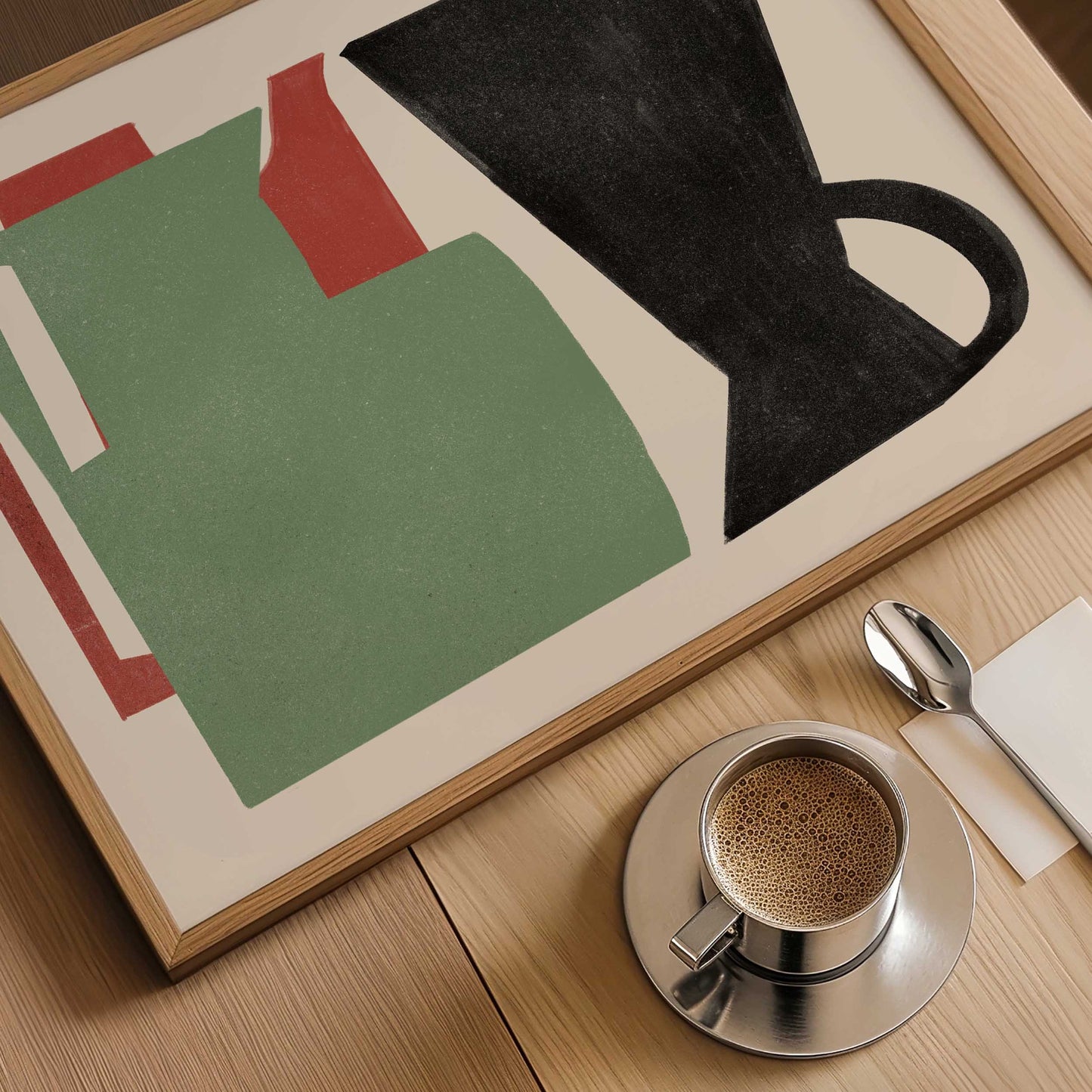 Framed abstract art print featuring a green shape, red accents, and a black coffee mug silhouette on a light wood table, styled with a cup of espresso, a spoon, and a napkin.
