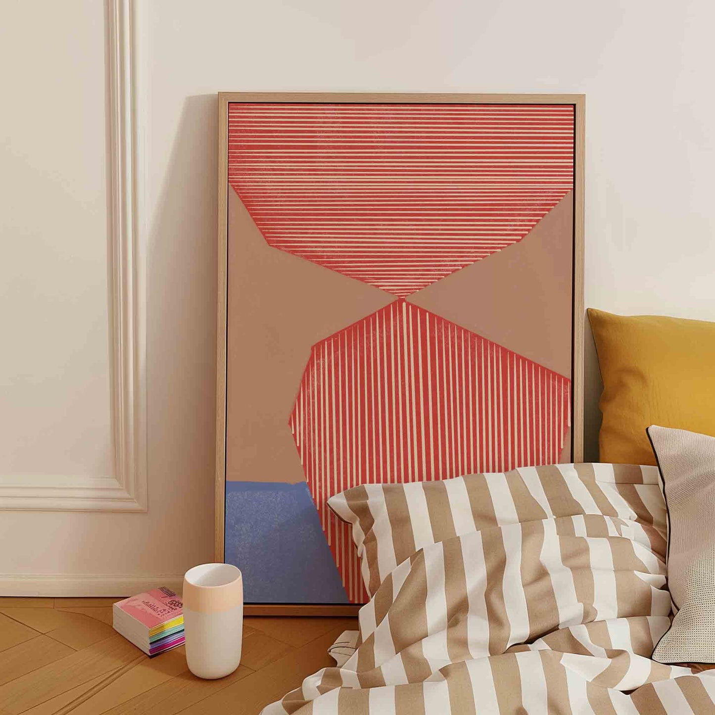 A framed abstract art print with coral, beige, and blue geometric shapes leans against a cream wall in a bedroom setting next to a bed with striped bedding.