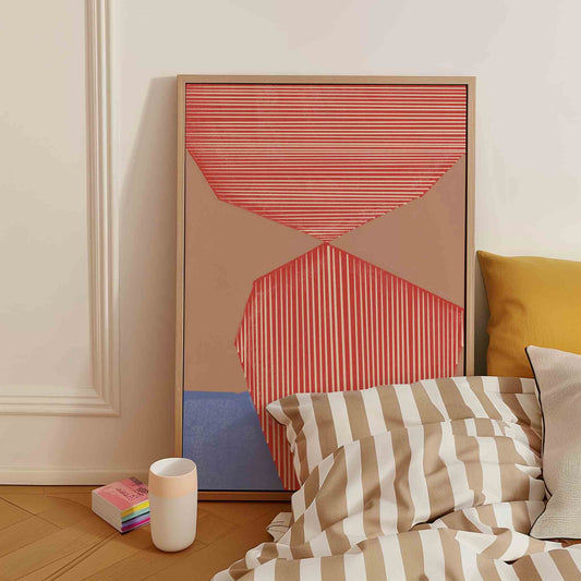 A framed abstract art print with coral, beige, and blue geometric shapes leans against a cream wall in a bedroom setting next to a bed with striped bedding.
