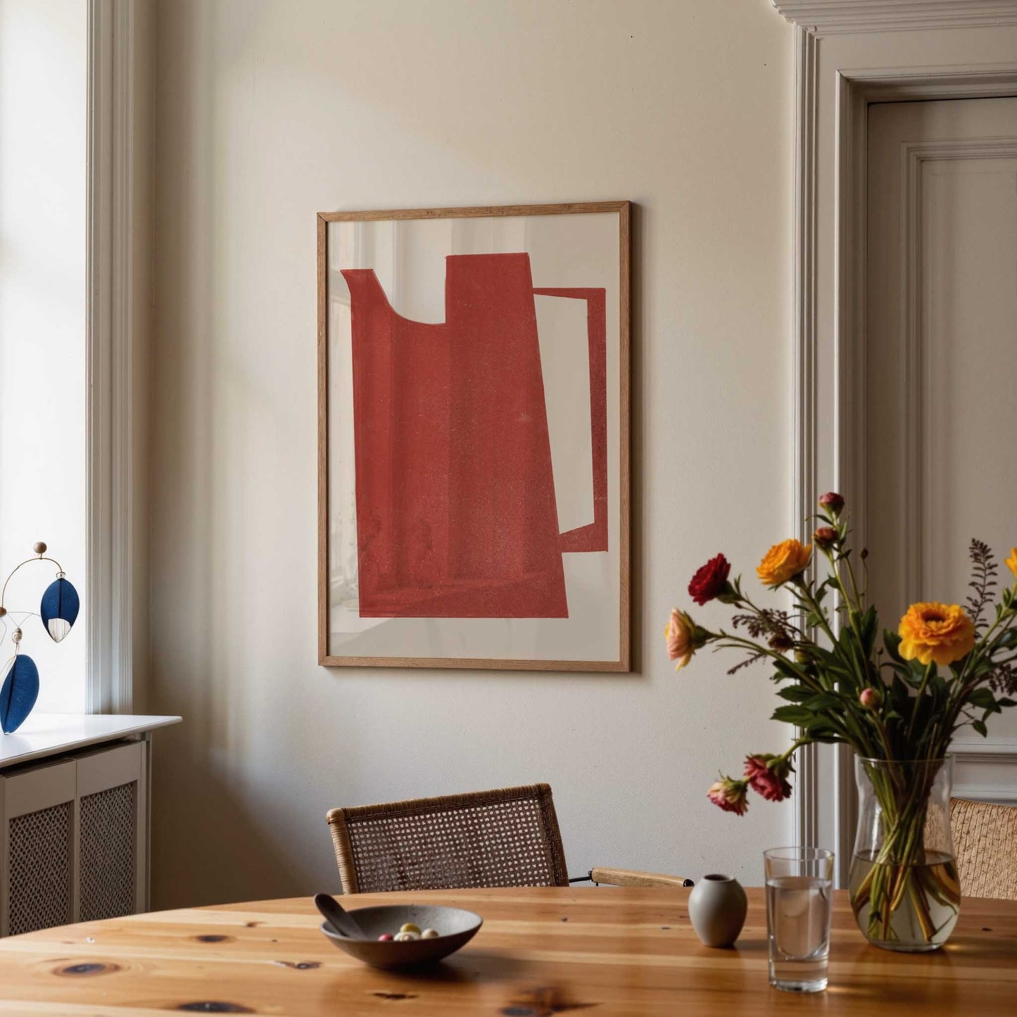Framed abstract wall art with a red geometric shape on a white background, hanging on a beige wall in a styled dining room with a wooden table, flowers, and a chair.