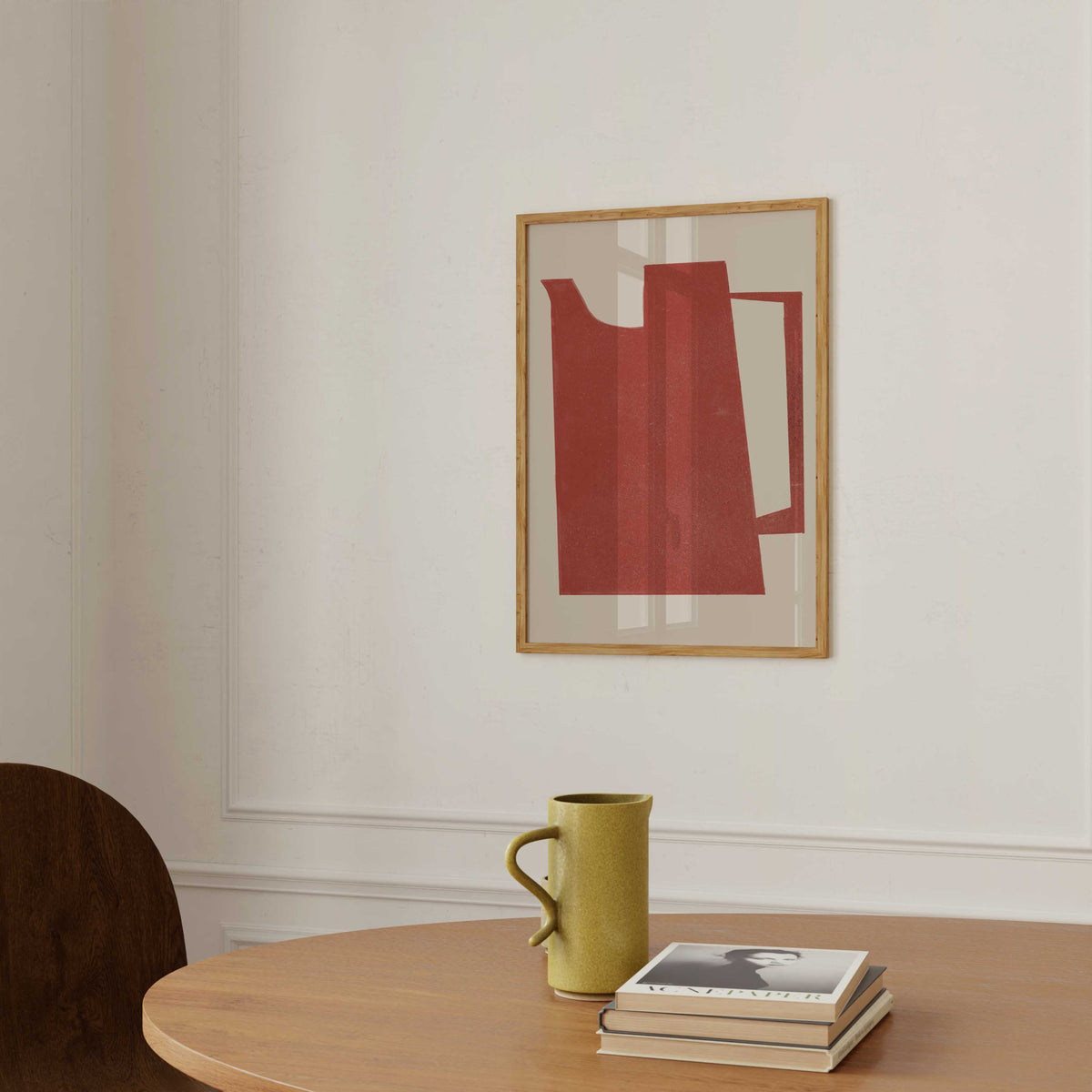 A framed abstract art print featuring a red geometric shape on a cream background, hanging on a white wall above a wooden table with a green mug and a stack of books.