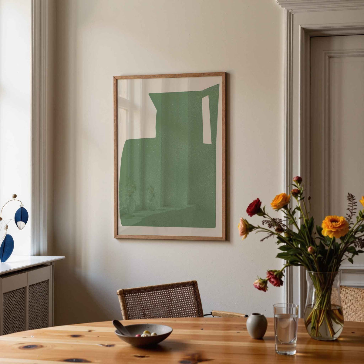 A framed abstract art print featuring a muted green geometric shape on a white background, hanging on a beige wall in a styled dining room setting with a wooden table, chair, and a vase of flowers.