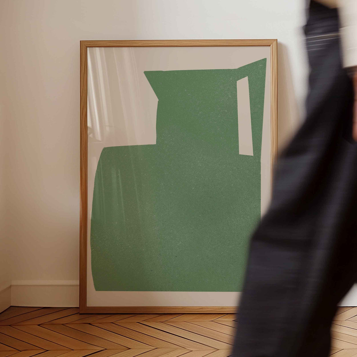 A framed abstract art print featuring a textured sage green shape with white negative spaces, set in a light wood frame and leaning against a cream wall on a wooden herringbone floor. A person in dark clothing is blurred in the foreground on the right.