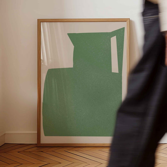 A framed abstract art print featuring a textured sage green shape with white negative spaces, set in a light wood frame and leaning against a cream wall on a wooden herringbone floor. A person in dark clothing is blurred in the foreground on the right.