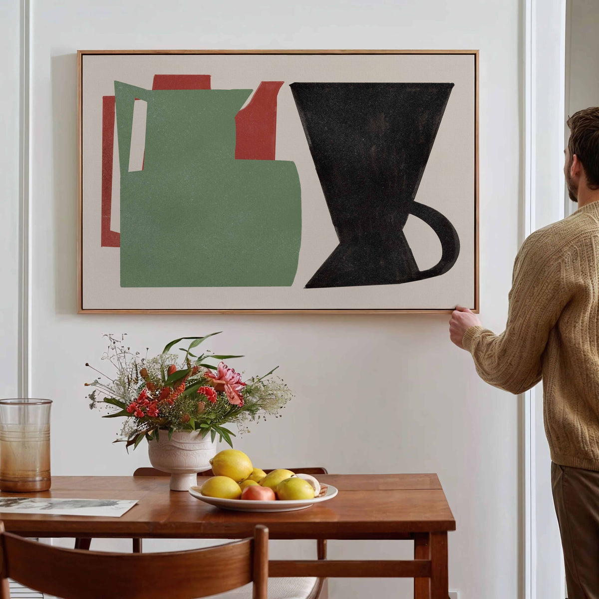 A man in a beige sweater hangs a framed abstract painting on a white wall above a wooden dining table with a vase of flowers and a plate of fruit. The painting features a green pitcher, a black coffee maker, and red geometric shapes on a cream background.