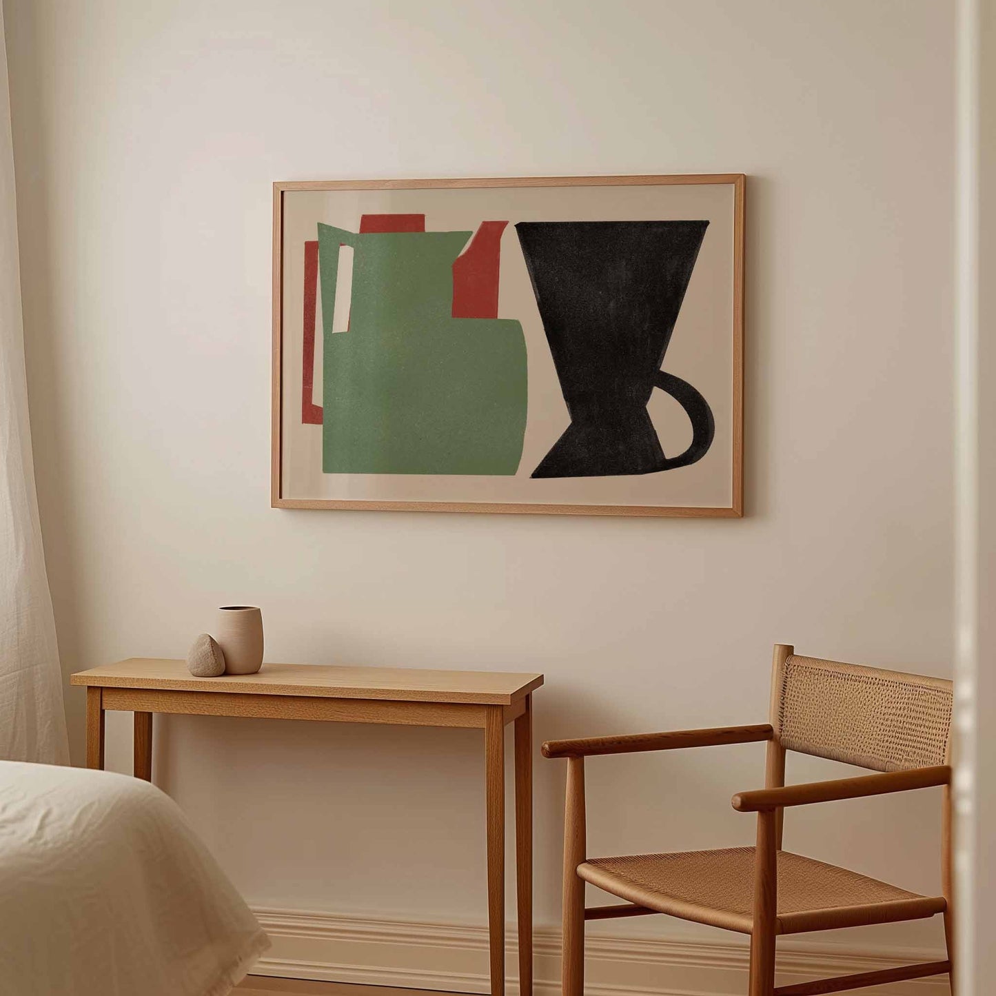 A framed abstract art print featuring a green pitcher, red accents, and a black coffee cup, displayed on a warm beige wall above a light wood console table and a woven-back wooden chair.